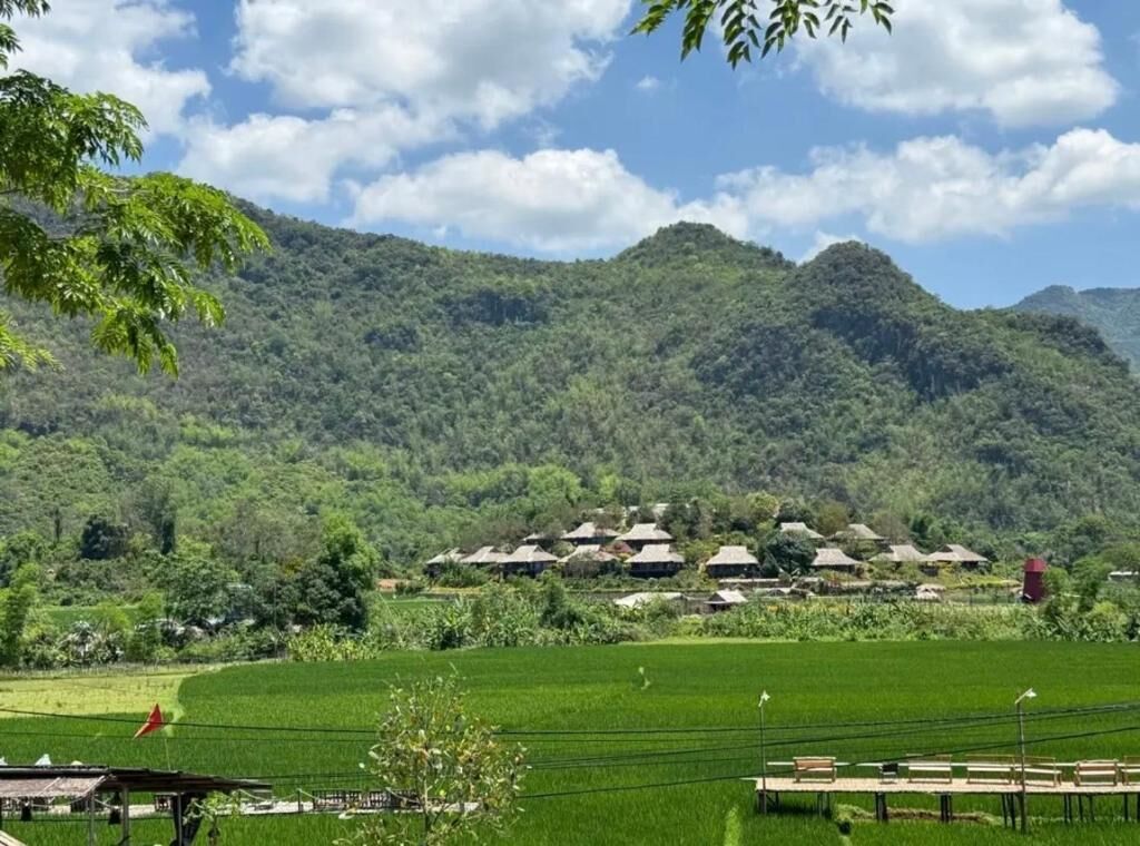 Ländliche Landschaft mit grünen Feldern, einer Gruppe traditioneller Häuser und einem Berg im Hintergrund.