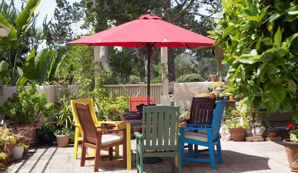 Un patio al aire libre con un paraguas rojo, sillas de madera y plantas en macetas.