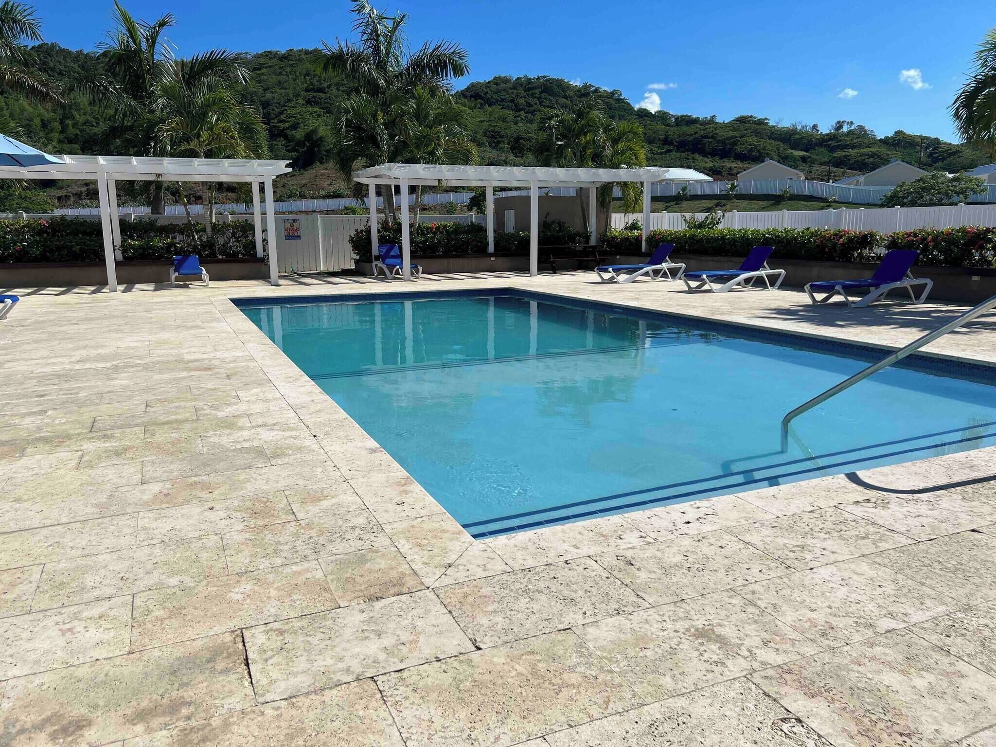 Uma piscina retangular com água azul cristalina, rodeada por um deck revestido com azulejos. Há cadeiras de descanso e uma área de estar coberta com um telhado branco.