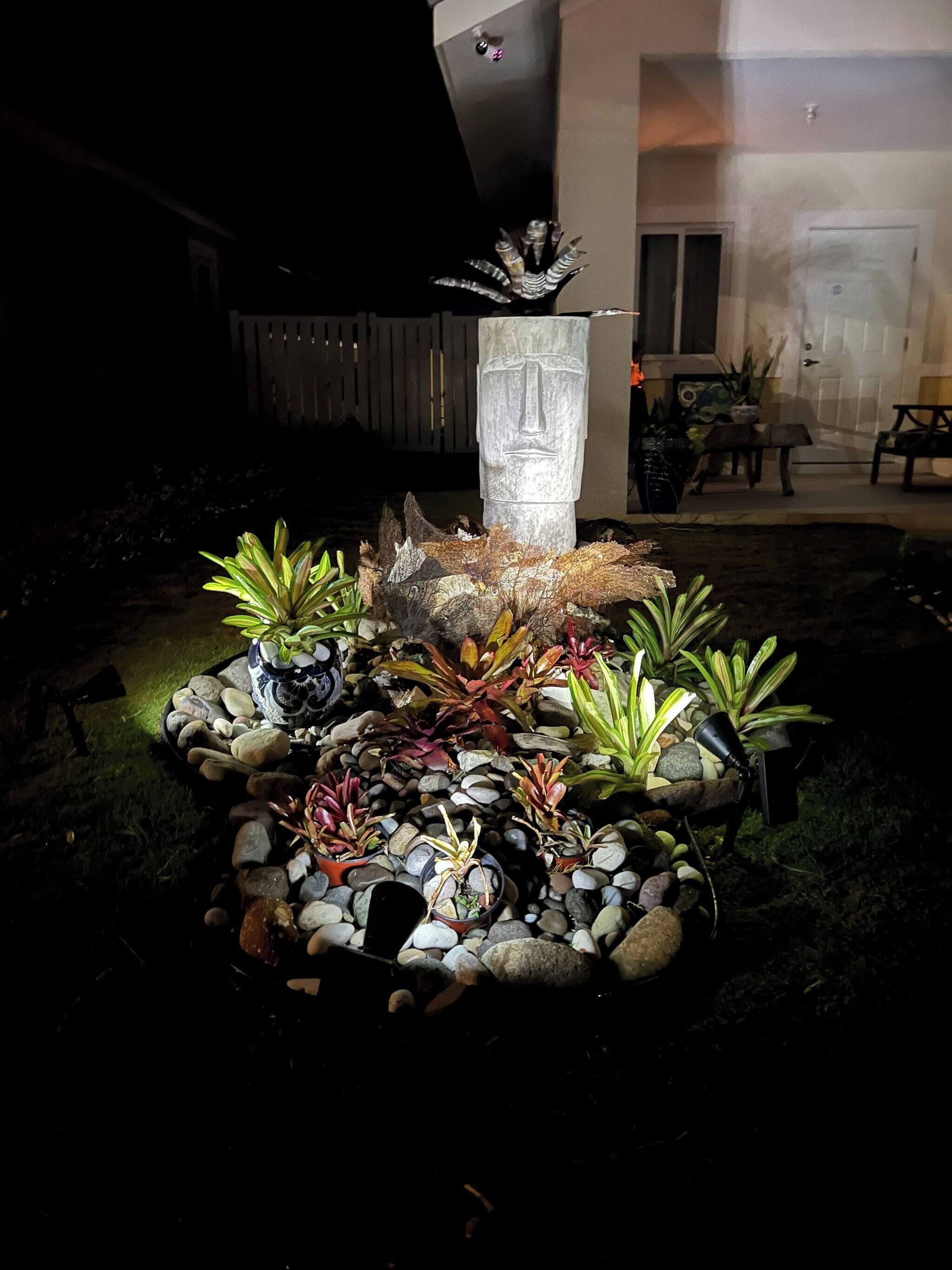 Um jardim com uma escultura de pedra, diversas plantas e um caminho de pedra.