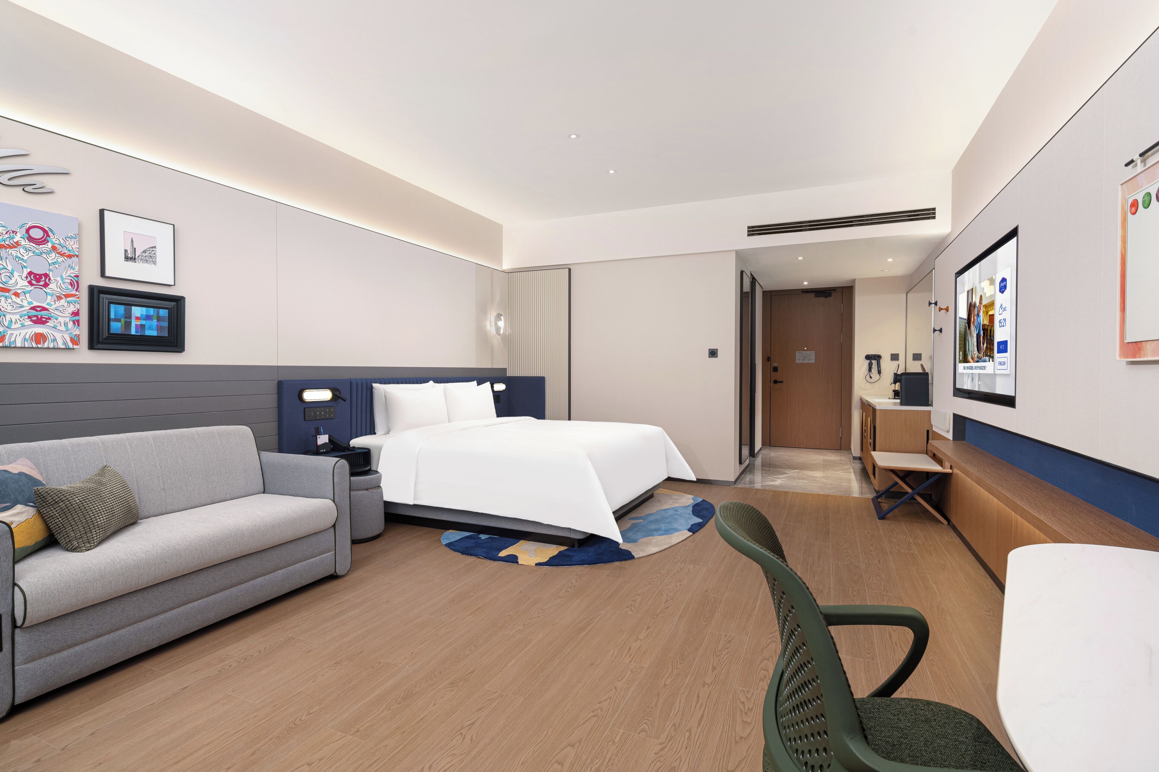 A hotel room with a bed, a sofa, a desk, and a chair. There are wall-mounted lights, a picture frame, and a small rug.