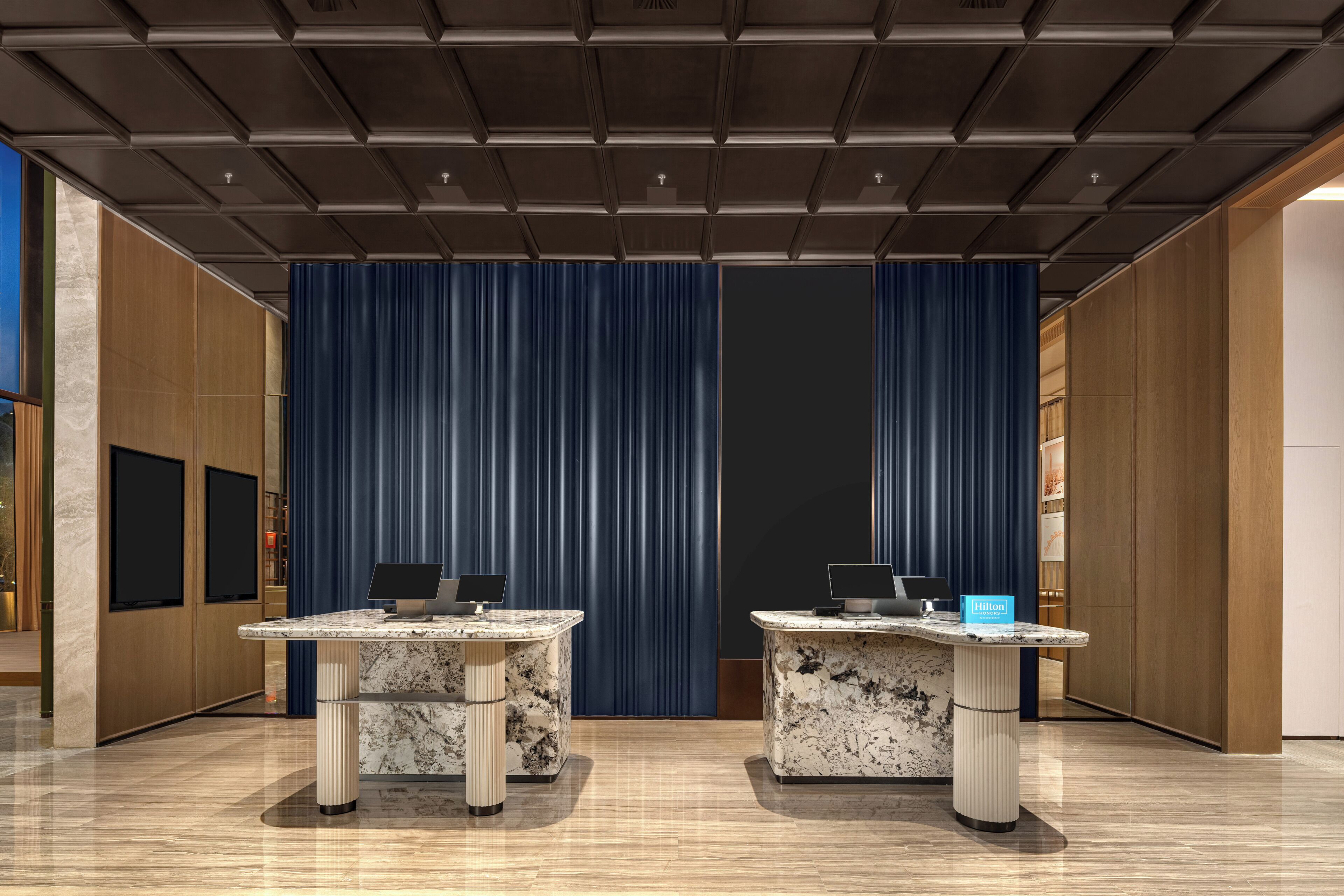 Vestíbulo de hotel con dos mostradores de recepción, un fondo de cortina azul y un diseño de techo moderno.