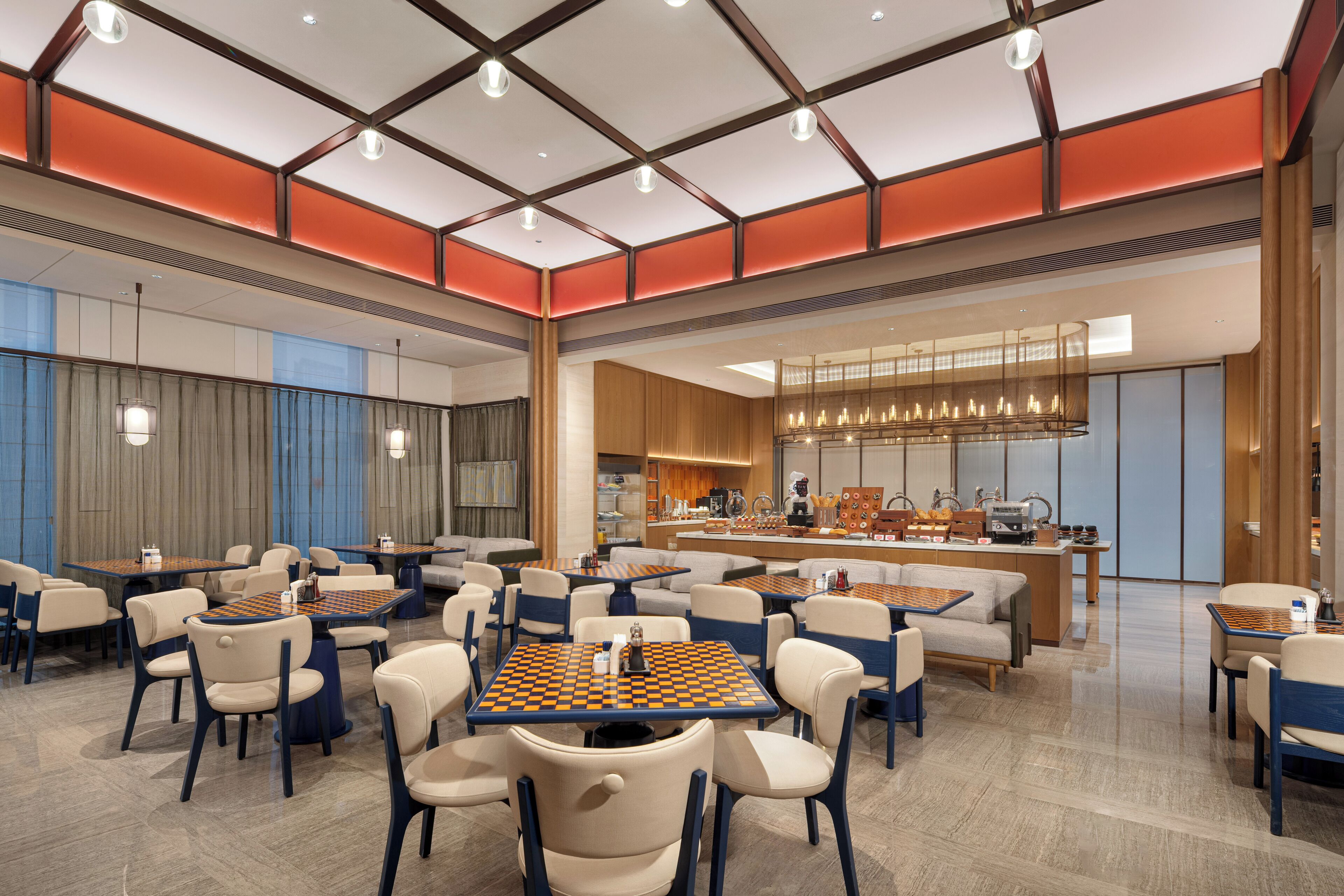 A modern hotel lobby with multiple seating areas, including tables and sofas, and a central bar area with a kitchen in the background.