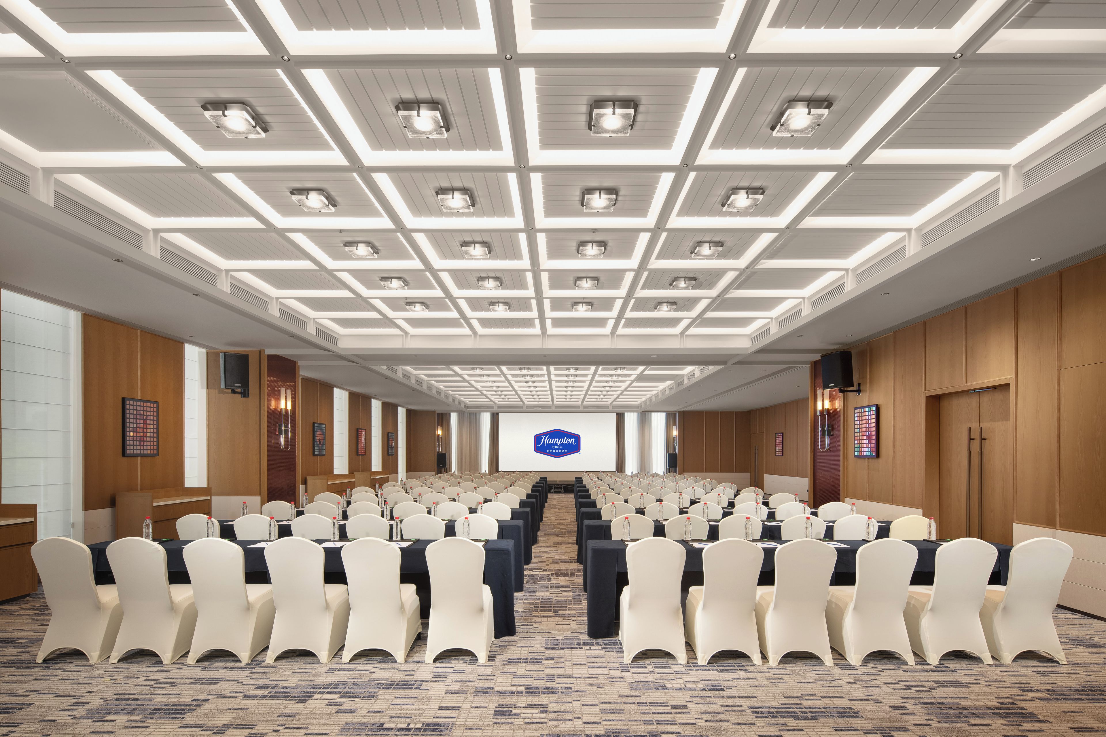 Een vergaderzaal met rijen stoelen, een groot scherm aan de voorkant en een plafond met inbouwverlichting.