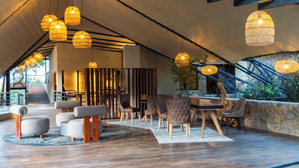A spacious indoor area with a high ceiling, wooden beams, and hanging pendant lights. There are several seating areas with cushioned chairs and a round table.