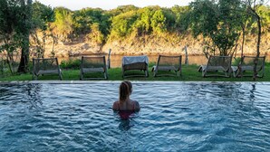 A pool with a person in the water, surrounded by lounge chairs and a natural landscape.