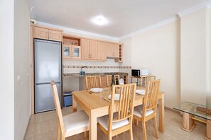 A compact kitchen with a refrigerator, wooden cabinets, a dining table with chairs, and a small glass table. - Charming 2-Bedroom Apartment in Playa San Juan, Spain (Guía de Isora)
