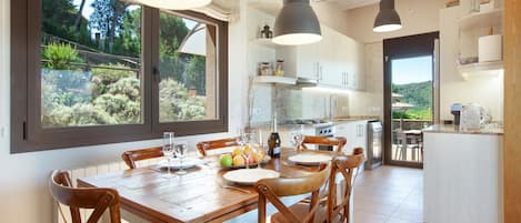 A dining area with a wooden table, chairs, and pendant lighting. There is a kitchen in the background with white cabinets and a view of greenery outside.