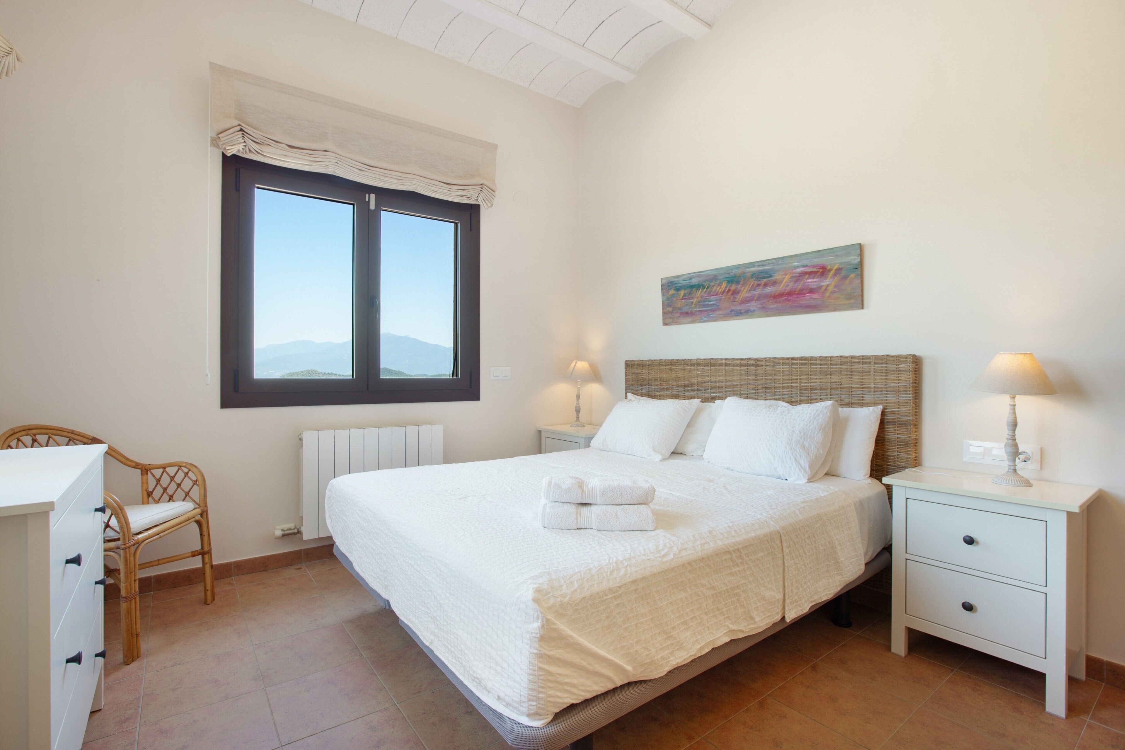 Un dormitorio con cama, mesitas de noche, una silla, una ventana y un cuadro en la pared.