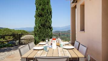 Mesa de madera al aire libre con sillas, una botella, vasos y un pastel en una terraza con vistas a las montañas.