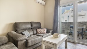Een woonkamer met een grijze bank, een klein wit tafeltje en uitzicht op gebouwen door een glazen deur.