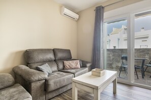 Ein Wohnzimmer mit grauem Sofa, einem kleinen weißen Tisch und Blick auf Gebäude durch eine Glastür.