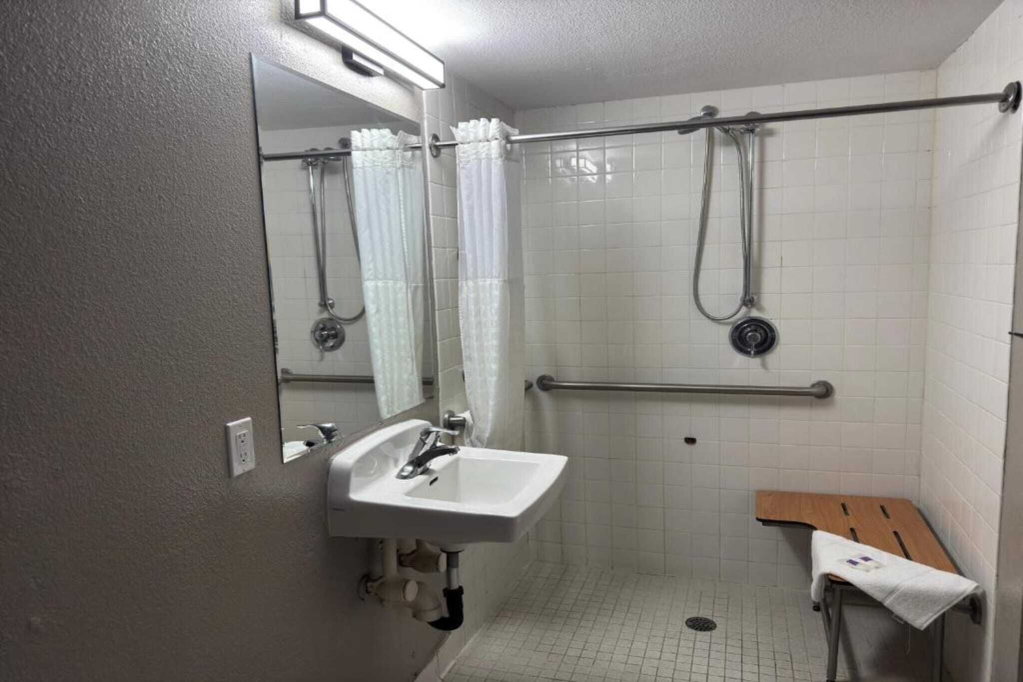A small, single-room bathroom with a white sink, a mirror, a shower with a grab bar, and a wooden bench.