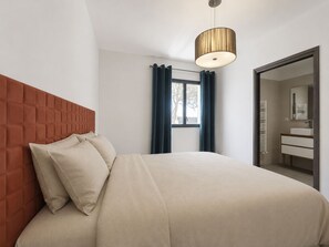A bedroom with a tufted headboard, a bed with beige bedding, a hanging light fixture, a window with curtains, and a bathroom visible through an open door. - E Follicie by Interhome (Miomo)