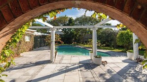 Une arche en briques mène à un espace piscine agrémenté d’une pergola et de plantes en pot.