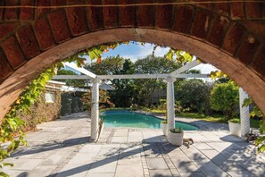 A brick archway leading to a swimming pool area with a pergola and potted plants.