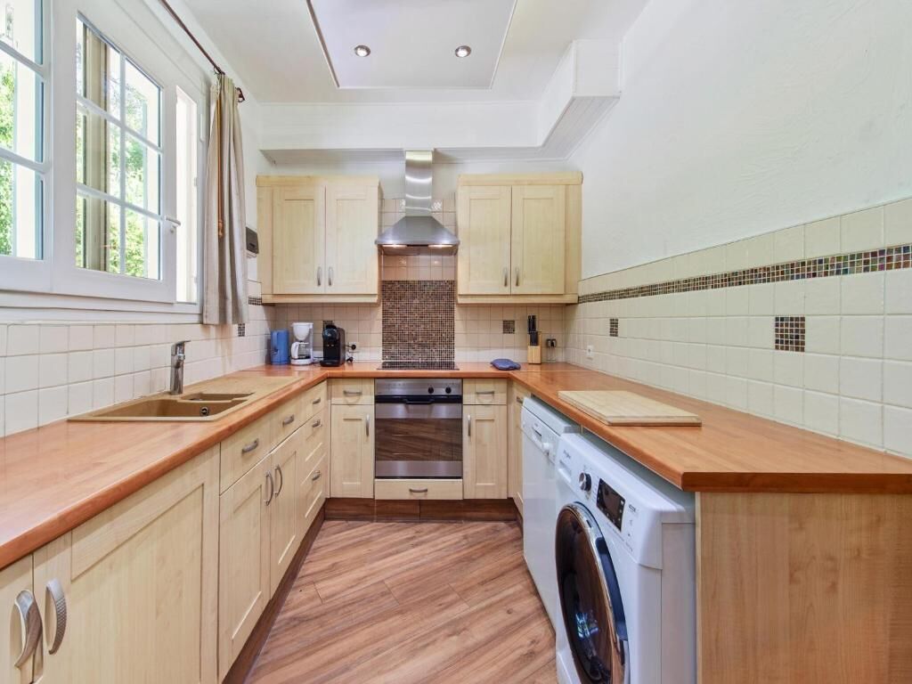 A kitchen with wooden cabinets, a stainless steel range hood, a built-in oven, a washing machine, and a large window.
