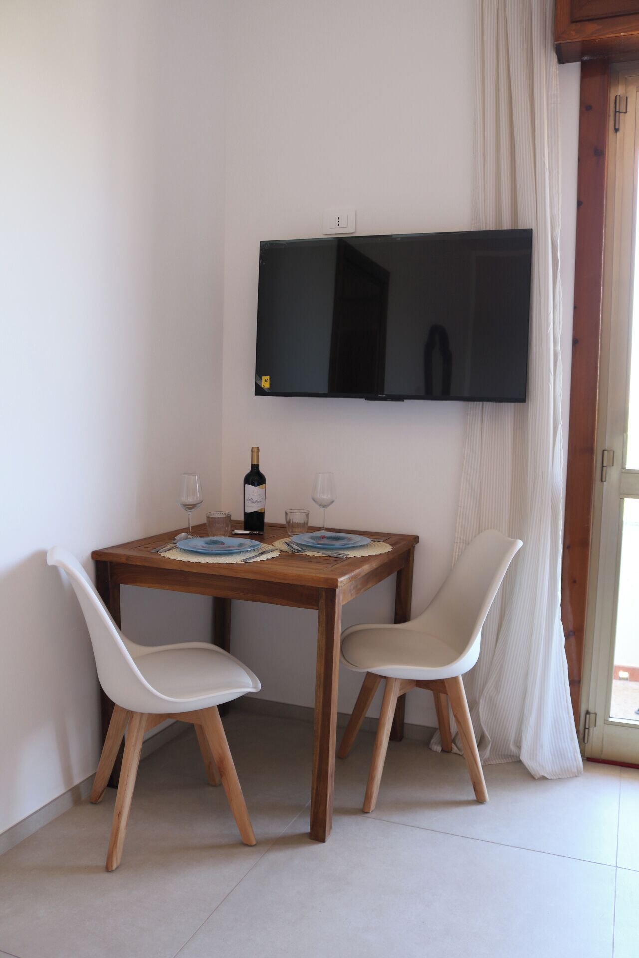 Un coin repas avec une table en bois, deux chaises, un téléviseur fixé au mur et une bouteille de vin.