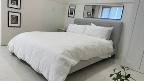 Una cama bien hecha con ropa blanca, un cabecero tapizado en gris y una mesita de noche pequeña.