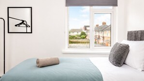 Ein Schlafzimmer mit einem Bett, einem Fenster mit Blick auf Häuser, einem gerahmten Bild und einem zusammengerollten Handtuch.