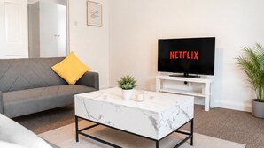 Ein Wohnzimmer mit einer Couch, einem Couchtisch, einem Fernseher, der 'NETFLIX' anzeigt, einer kleinen Pflanze und einem gerahmten Zitat an der Wand.