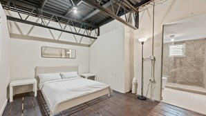 Ein Schlafzimmer mit einem Bett, Nachttischen, einer Bank, einer Stehlampe und einem Fenster mit Jalousien.