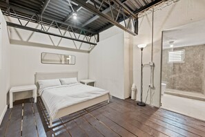 Ein Schlafzimmer mit einem Bett, Nachttischen, einer Bank, einer Stehlampe und einem Fenster mit Jalousien.