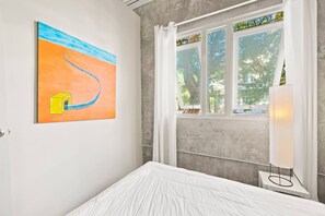 Un dormitori amb un llit, un quadre a la paret, una finestra amb cortines i una làmpada.