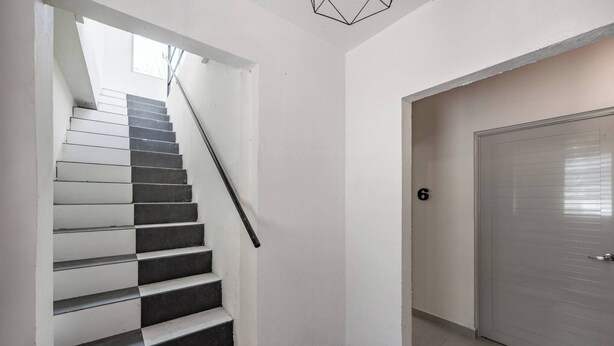 Eine Treppe mit abwechselnd schwarzen und weißen Stufen führt zu einer Zwischenebene, wo sich eine Tür mit der Nummer 6 befindet, und an der Decke hängt ein geometrisches Lichtobjekt.