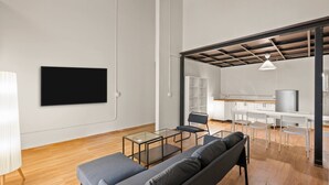 Ein modernes Wohnzimmer mit einer Couch, einem Glastisch, einem an der Wand befestigten Fernseher und einem durch eine offene Tür einsehbaren Küchenbereich.