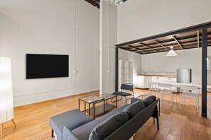 Ein modernes Wohnzimmer mit einer Couch, einem Glastisch, einem an der Wand befestigten Fernseher und einem durch eine offene Tür einsehbaren Küchenbereich.