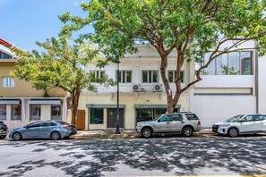 Une vue de rue avec des voitures garées, un immeuble doté de grandes fenêtres et un arbre qui offre de l’ombre.