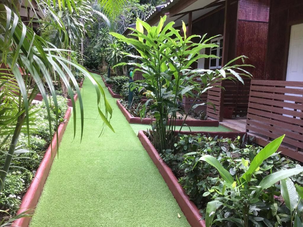 Uma passarela com grama sintética, rodeada por plantas diversas e um banco de madeira.
