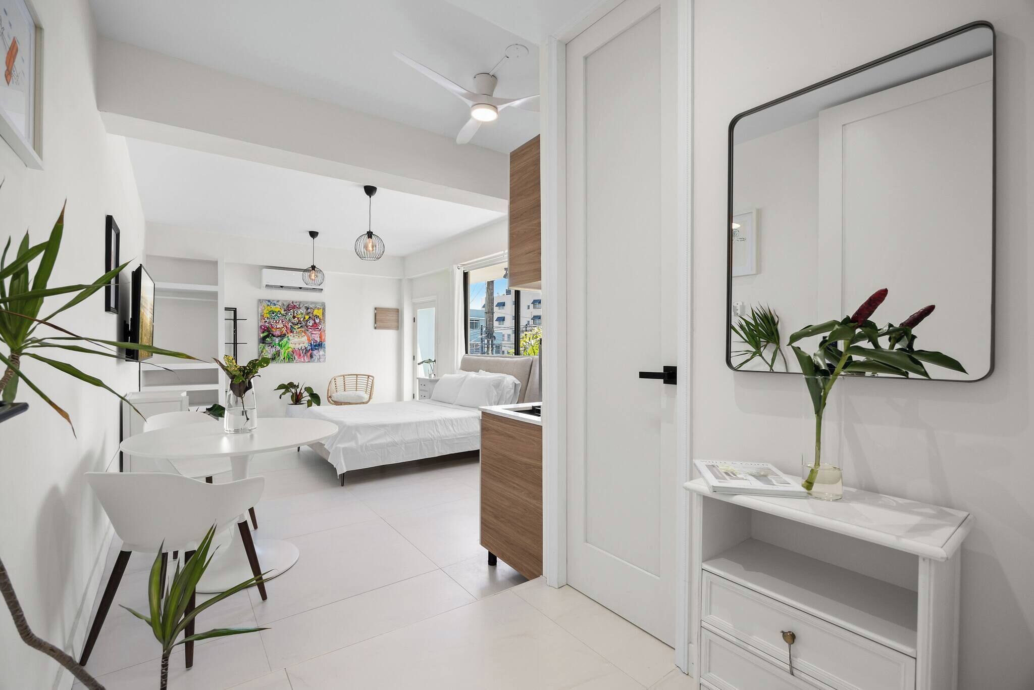 A modern, well-lit room with a bed, a dining table with chairs, a white TV, and a large mirror.
