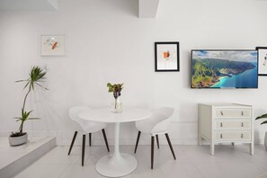 Ein modernes Zimmer mit einem runden weißen Tisch, zwei weißen Stühlen, einer kleinen weißen Kommode und gerahmten Kunstwerken an der Wand.