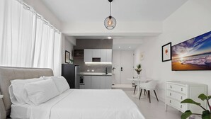 Un dormitorio moderno con cama, escritorio, silla, una pequeña kitchenette y un ventanal con cortinas.