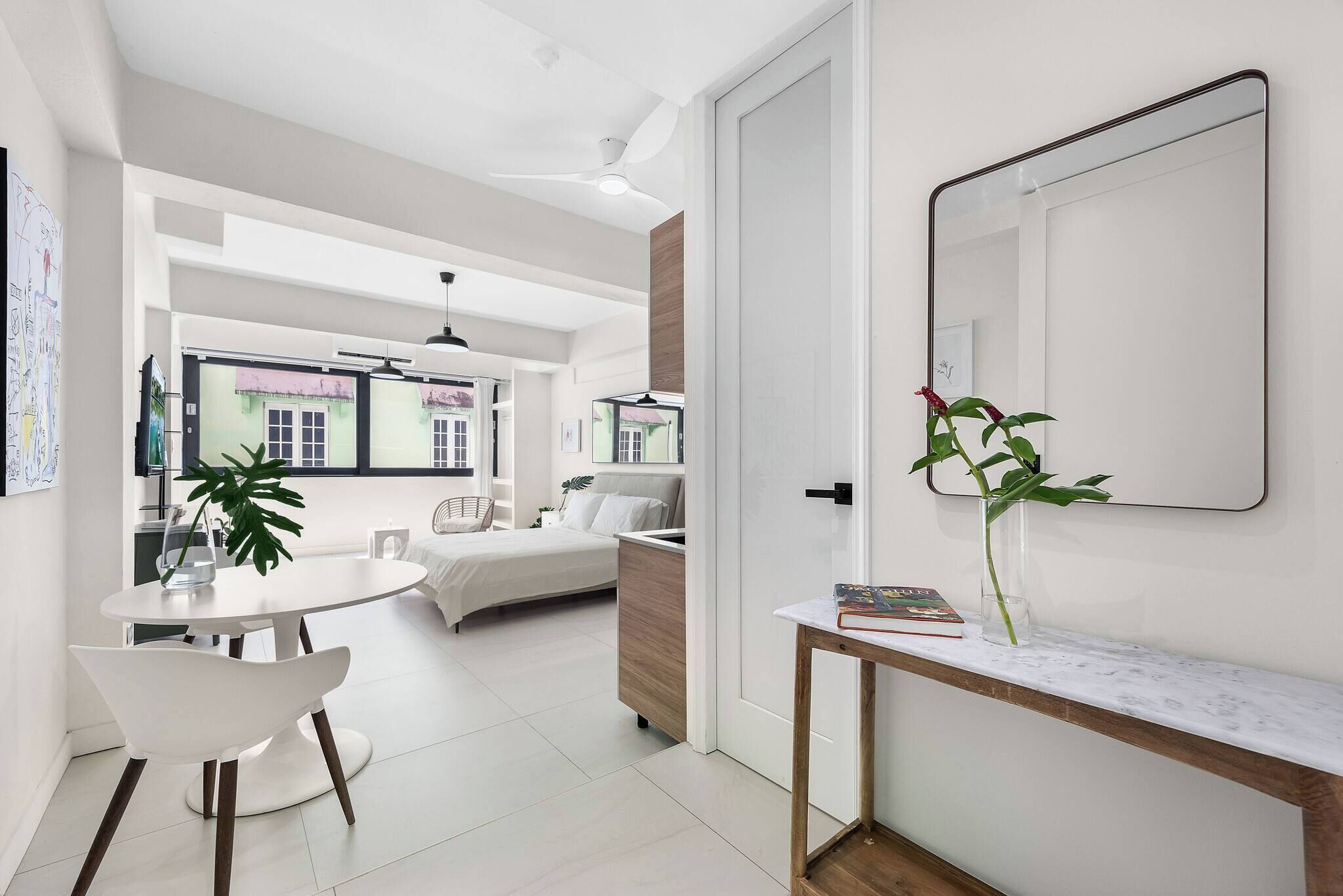 A modern, well-lit room with a dining area, a bed, a large mirror, and a potted plant.