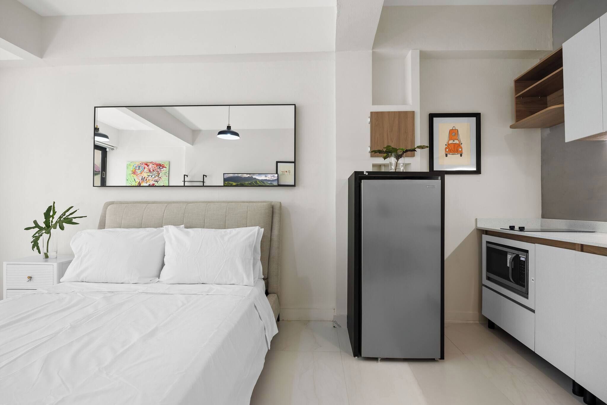 A modern bedroom with a bed, a bedside table with a plant, a large mirror, and a kitchen area with a refrigerator and microwave.