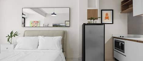 A modern bedroom with a bed, a bedside table with a plant, a large mirror, and a kitchen area with a refrigerator and microwave.
