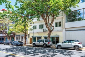 Straßenansicht mit parkenden Autos, einem Baum und einem Gebäude mit weißer Fassade und grünen Fenstern.
