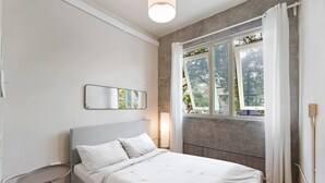 A bedroom with a bed, a bedside table, a mirror, a window with curtains, and a view of greenery outside. - Barrio Sanctuary - Luna (San Juan city)