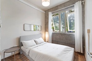 Ein Schlafzimmer mit Bett, Nachttisch, Spiegel, einem Fenster mit Vorhängen und Blick ins Grüne.