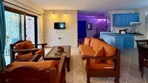 Ein Wohnzimmer mit Sofa, Sesseln, einem Couchtisch, einem an einer strukturierten Wand montierten Fernseher und einer Küchenzeile mit blauen Schränken und einem Herd.