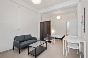 A modern room with a sofa, a glass-top table, a white chair, a bed, and a desk with a mirror. - Barrio Sanctuary - Encanto (San Juan city)