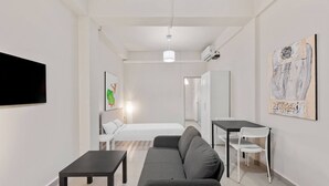 Ein modernes, gut beleuchtetes Zimmer mit grauem Sofa, einem kleinen Tisch, einem Bett und einem Schreibtisch mit Stühlen.