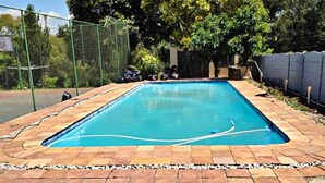 Et rektangulært swimmingpool med flisebelagt terrasse, omgivet af hegn og træer.