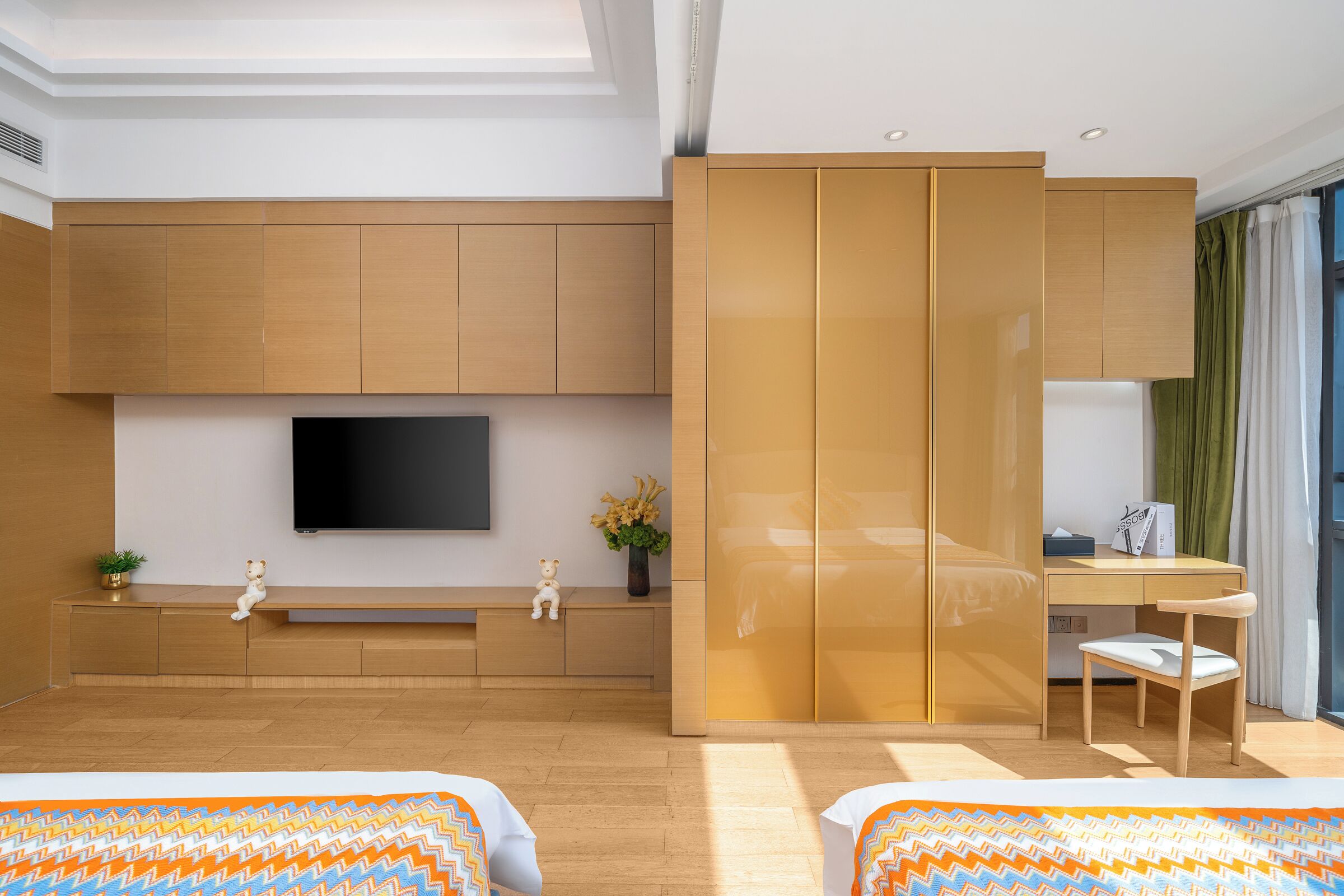 A modern hotel room with a large flat-screen TV, wooden cabinetry, a desk with a chair, and a bed with patterned bedding.