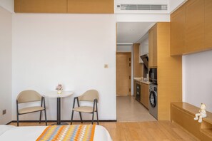 Habitación de hotel con cama, dos sillones, una mesita con un florero, una lavadora al fondo y una figura de oso en un banco.