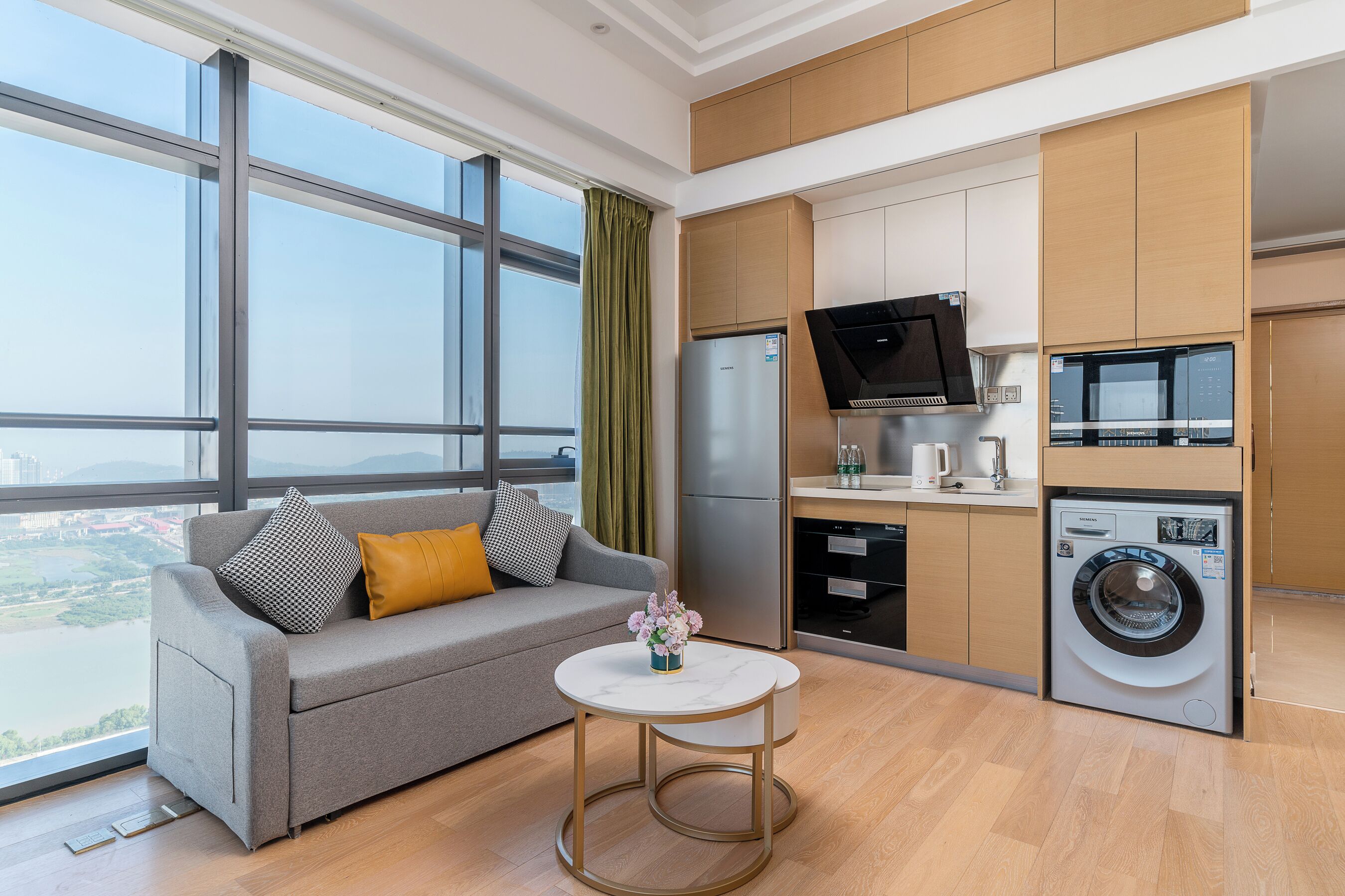 A modern kitchen with a built-in washer and dryer, a grey sofa with cushions, and a small round table with a vase.