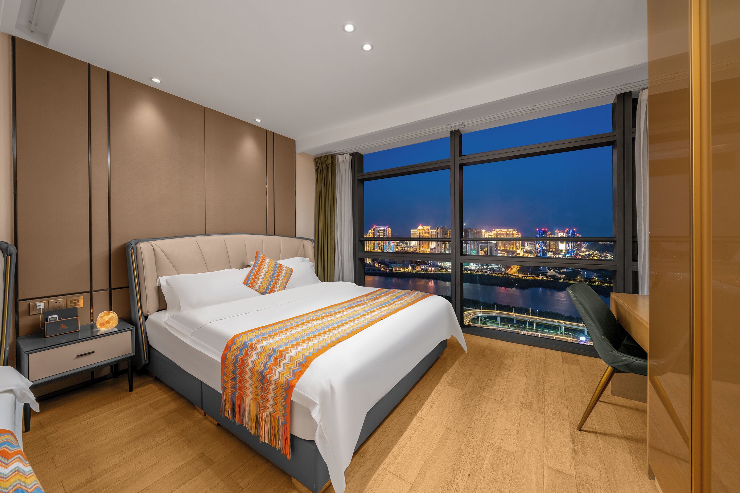 A modern hotel room with a large bed, a nightstand, a chair, and a city view.